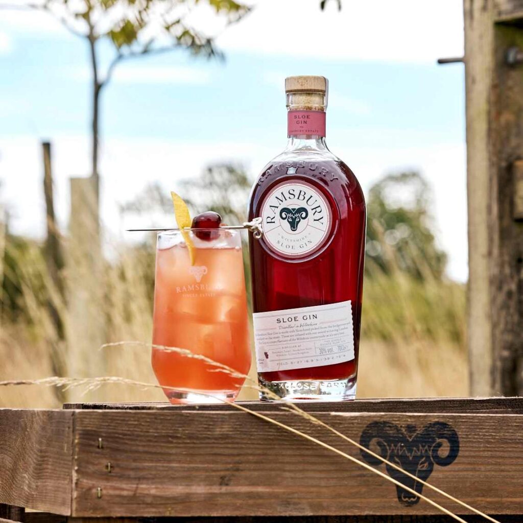 A bottle of Ramsbury Sloe Gin alongside a filled cocktail glass garnished with a lemon twist and a berry on a cocktail pick, set outdoors on a wooden crate.