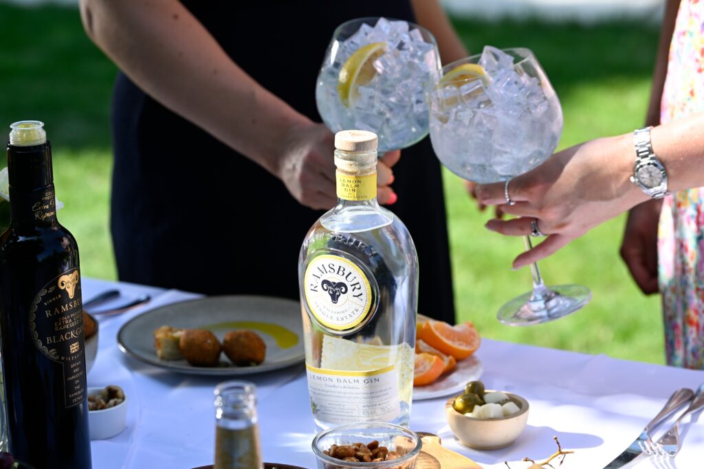 A bottle of Ramsbury Lemon Balm Gin on an outdoor table surrounded by dishes, as two people toast with gin and tonic glasses filled with ice and lemon slices.